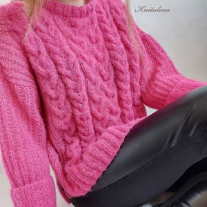 Op de afbeelding: Een roze, pluche kabelgebreide trui met lange mouwen en een ronde hals. De trui wordt gedragen door een persoon met een zwarte leren broek en witte sokken met een sterrenpatroon. De tekst "Knitalina" is zichtbaar in de rechterbovenhoek van de afbeelding.