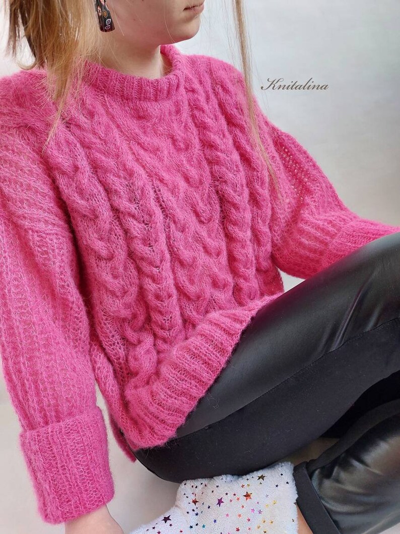 Op de afbeelding: Een roze, pluche kabelgebreide trui met lange mouwen en een ronde hals. De trui wordt gedragen door een persoon met een zwarte leren broek en witte sokken met een sterrenpatroon. De tekst "Knitalina" is zichtbaar in de rechterbovenhoek van de afbeelding.