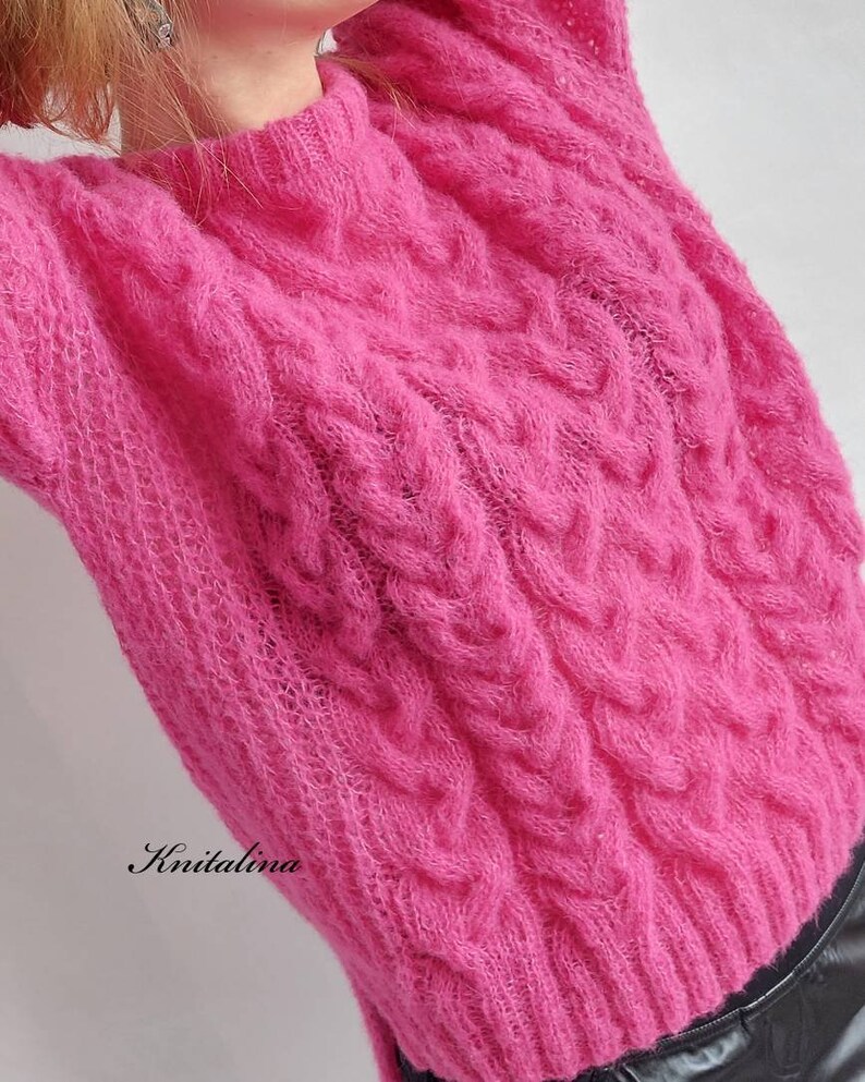 Op de afbeelding: Een roze, pluche kabelgebreide trui met een ronde hals. De trui heeft een losse pasvorm en is gemaakt van een zacht, comfortabel garen.