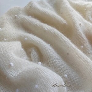 May include: A white knitted shawl with a delicate, open weave and small white beads throughout. The shawl is soft and fluffy, with a subtle sheen.