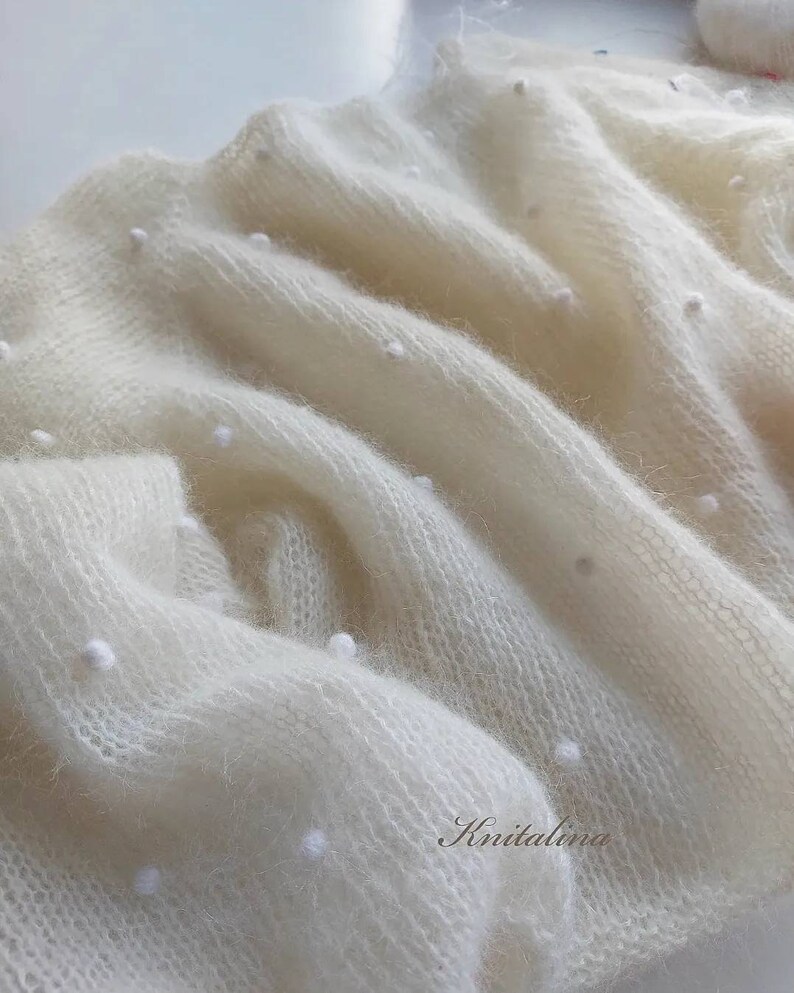 May include: A white knitted shawl with a delicate, open weave and small white beads throughout. The shawl is soft and fluffy, with a subtle sheen.