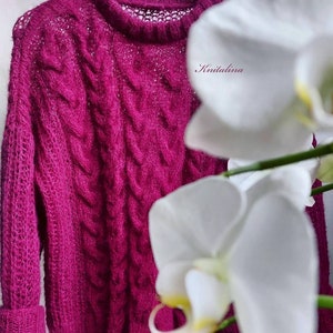 Op de afbeelding: Een magenta gebreide trui met een kabelbreipatroon en een delicate opengewerkte halslijn. De trui hangt aan een witte hanger en wordt gedeeltelijk bedekt door een witte orchidee.