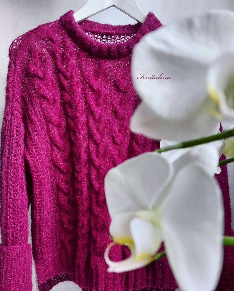 Op de afbeelding: Een magenta gebreide trui met een kabelbreipatroon en een delicate opengewerkte halslijn. De trui hangt aan een witte hanger en wordt gedeeltelijk bedekt door een witte orchidee.