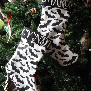 May include: Two black and white Christmas stockings with a bat pattern. The stockings are personalised with the names "Sharon" and "Cyndi".
