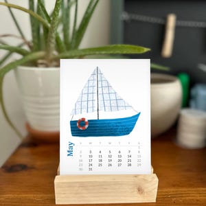May include: A desk calendar page for May featuring a blue sailboat illustration with a white checkered sail and a red and white life preserver. The calendar grid displays the days of the month below the sailboat graphic on a white background.