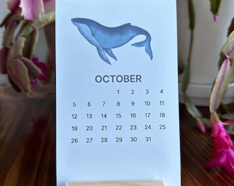 May include: A white paper calendar page for October featuring a blue whale illustration at the top. The calendar grid displays the numbers 1 through 31 in black text. The page is held upright by a rectangular light-colored wooden block stand.