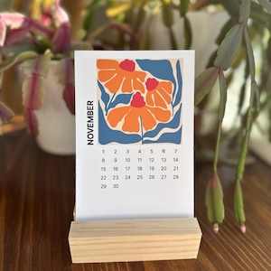 Può includere: Una pagina di calendario in carta bianca per novembre con un'illustrazione grafica di tre fiori arancioni con centri rossi e foglie blu. La griglia del calendario elenca le date dall'1 al 30, sostenuta da un supporto in legno di colore chiaro.