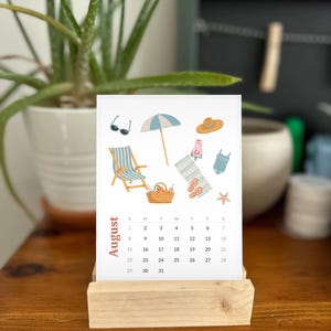 May include: A white paper calendar page for August displayed on a light wood stand. The top section features colorful illustrations of beach items including a striped chair, umbrella, hat, sunglasses, swimsuit, sandals, and a picnic basket.