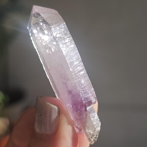 Op de afbeelding: Een helder kwartskristal met een paars amethistcentrum. Het kristal wordt in het licht gehouden, waardoor de facetten en de natuurlijke textuur zichtbaar worden. Het kristal heeft een lichtpaarse kleur met een heldere punt.