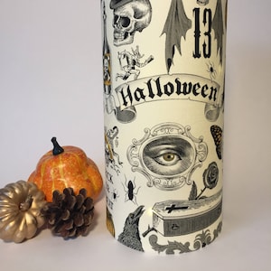 Könnte beinhalten: Ein zylindrischer Halloween-Lampenschirm mit cremefarbenem Hintergrund und schwarz-weißen Illustrationen von Totenköpfen, Fledermäusen und anderen gruseligen Elementen. Das Wort "Halloween" ist auf einem Banner zu sehen. Ein kleiner Kürbis und ein Tannenzapfen befinden sich neben der Lampe.