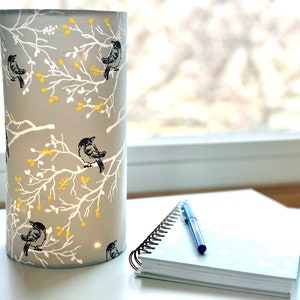 May include: A cylindrical lamp shade featuring a light gray background with a white branch pattern, small yellow leaves, and black birds perched on the branches. The lamp shade is illuminated from within, casting a soft glow through the fabric.