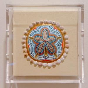 以下が含まれることがあります： 青、オレンジ、白の複雑な模様が描かれたサンドダラーが、透明なアクリルボックスに収められています。サンドダラーの周囲は小さな明るい色の貝殻で縁取られ、正方形のクリーム色の布製キャンバスに固定されています。