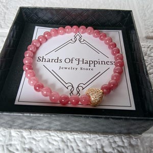 May include: A pink beaded bracelet with a gold heart charm, presented in a black box. The bracelet is made of small, round, pink beads. The box contains a card with the text "Shards Of Happiness Jewelry Store."