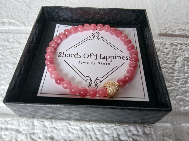 May include: A pink beaded bracelet with a gold heart charm, presented in a black box. The bracelet is made of small, round, pink beads. The box contains a card with the text "Shards Of Happiness Jewelry Store."