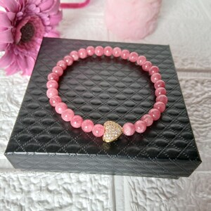May include: A pink beaded bracelet with a gold heart-shaped charm, resting on a black box. The bracelet is made of small, round, pink beads. The heart charm is encrusted with small, sparkling stones. A pink flower is visible in the background.