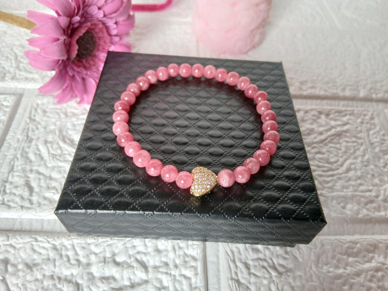 May include: A pink beaded bracelet with a gold heart-shaped charm, resting on a black box. The bracelet is made of small, round, pink beads. The heart charm is encrusted with small, sparkling stones. A pink flower is visible in the background.