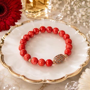 May include: A beaded bracelet featuring round, textured red stones and small gold-toned spacers. The bracelet includes a single, larger oval-shaped gold-toned bead with intricate filigree detailing and small embedded turquoise and black stones.