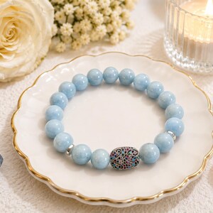 May include: A beaded bracelet featuring smooth, round, light blue gemstone beads and a single oval-shaped focal bead encrusted with small, multicolored crystals. The bracelet includes small silver-toned metal spacers between the beads.