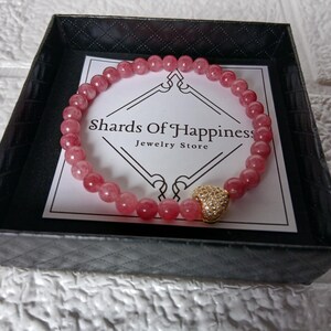 May include: A pink beaded bracelet with a gold heart-shaped charm, presented in a black box with a white label that reads "Shards Of Happiness Jewelry Store". The bracelet is made of small, round, pink beads.
