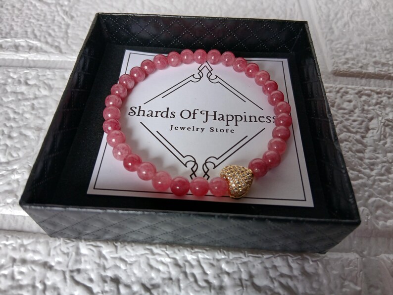 May include: A pink beaded bracelet with a gold heart-shaped charm, presented in a black box with a white label that reads "Shards Of Happiness Jewelry Store". The bracelet is made of small, round, pink beads.