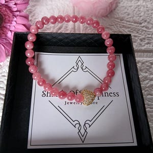 May include: A pink gemstone bracelet with a gold heart-shaped charm, presented in a black box. The bracelet is made of small, round, pink beads. The charm is encrusted with small, clear stones. The box has a white card with text.