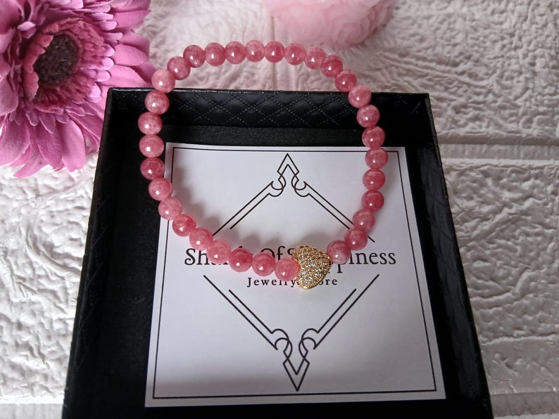 May include: A pink gemstone bracelet with a gold heart-shaped charm, presented in a black box. The bracelet is made of small, round, pink beads. The charm is encrusted with small, clear stones. The box has a white card with text.