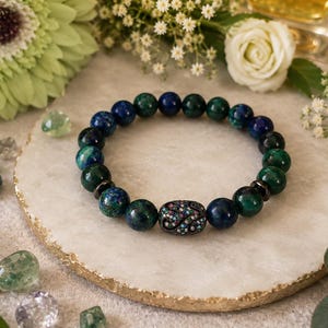 May include: A beaded bracelet featuring round, polished azurite and malachite stones in shades of deep blue and forest green. The bracelet includes a central oval-shaped metal bead encrusted with small, multicolored crystals and two dark spacers.