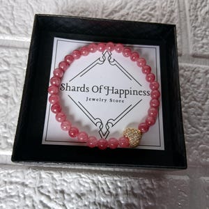 May include: A pink beaded bracelet with a gold heart charm, presented in a black box. The bracelet is made of small, round, pink beads. The heart charm is covered in small, sparkling stones. The box contains a white card with the text "Shards Of Happiness Jewelry Store".