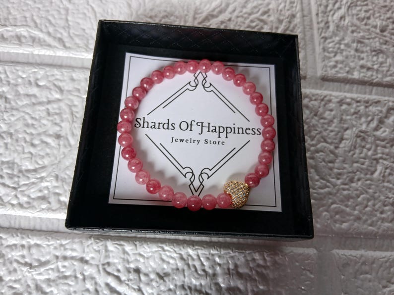 May include: A pink beaded bracelet with a gold heart charm, presented in a black box. The bracelet is made of small, round, pink beads. The heart charm is covered in small, sparkling stones. The box contains a white card with the text "Shards Of Happiness Jewelry Store".