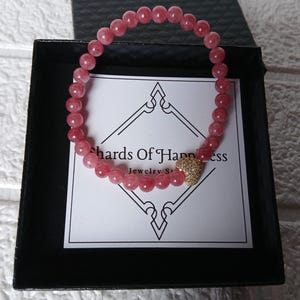 May include: A pink beaded bracelet with a gold heart-shaped charm, presented in a black box. The bracelet is made of small, round, pink beads. The box contains a card with the text "Shards Of Happiness Jewelry Store".