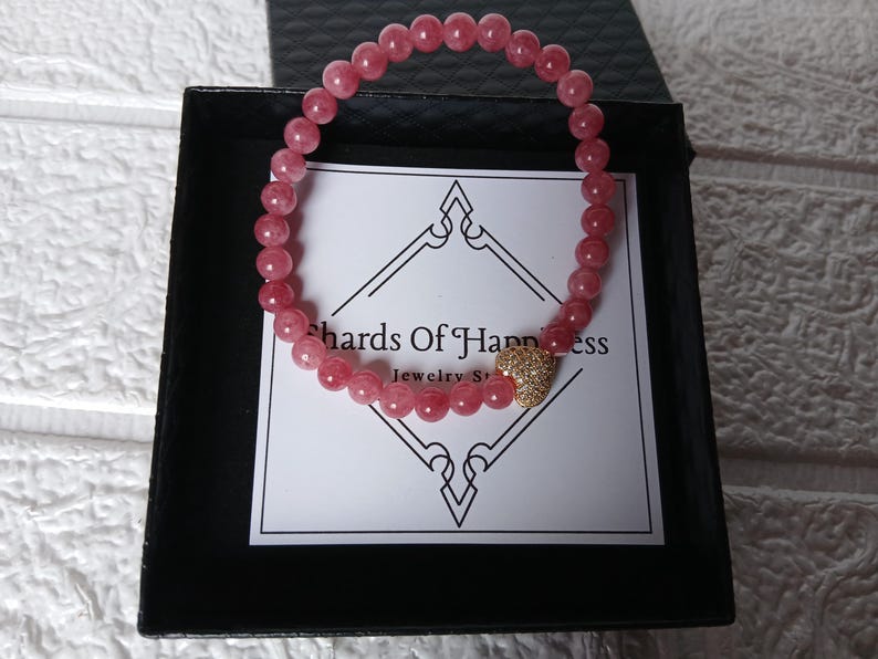 May include: A pink beaded bracelet with a gold heart-shaped charm, presented in a black box. The bracelet is made of small, round, pink beads. The box contains a card with the text "Shards Of Happiness Jewelry Store".