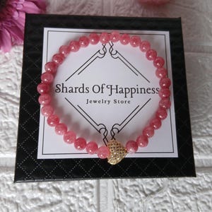 May include: A pink beaded bracelet with a gold-colored heart-shaped charm. The bracelet is displayed on a black box with the text "Shards Of Happiness Jewelry Store". A pink flower is visible in the background.