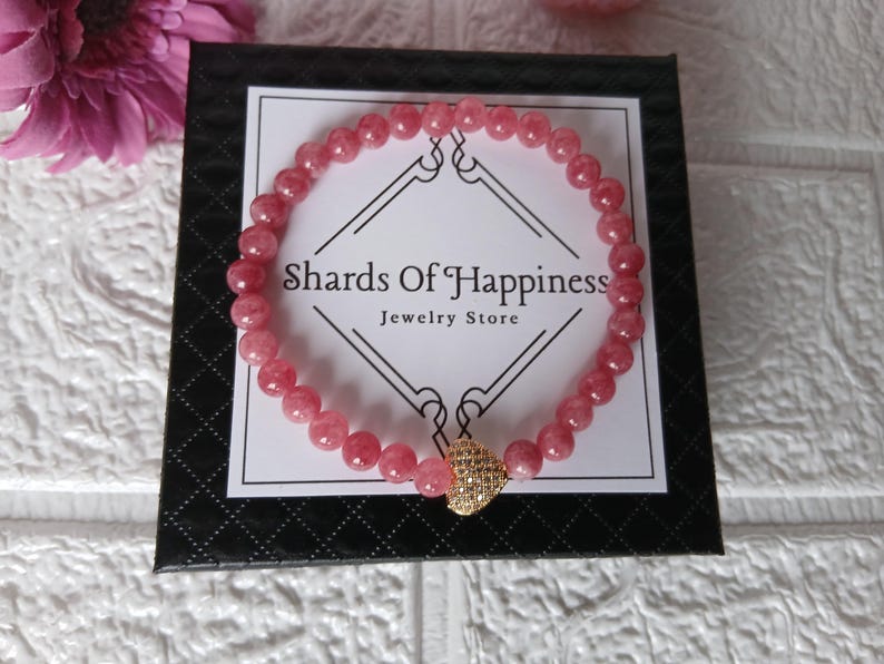 May include: A pink beaded bracelet with a gold-colored heart-shaped charm. The bracelet is displayed on a black box with the text "Shards Of Happiness Jewelry Store". A pink flower is visible in the background.