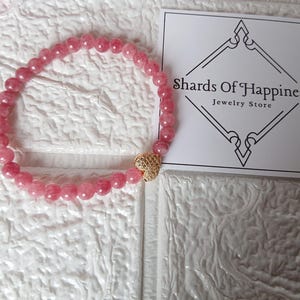May include: A pink beaded bracelet with a gold heart-shaped charm. The bracelet is made of small, round, pink beads. The charm is gold and has a textured surface. A white card with the text "Shards Of Happiness Jewelry Store" is in the background.