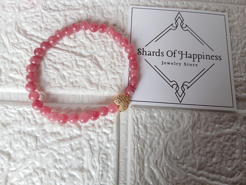 May include: A pink beaded bracelet with a gold heart-shaped charm. The bracelet is made of small, round, pink beads. The charm is gold and has a textured surface. A white card with the text "Shards Of Happiness Jewelry Store" is in the background.