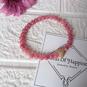 May include: A pink beaded bracelet with a gold heart charm. The bracelet is made of small, round, pink beads. The charm is gold and encrusted with small, clear stones. A card with the text "Status Of Happiness Jewelry Store" is also in the image.