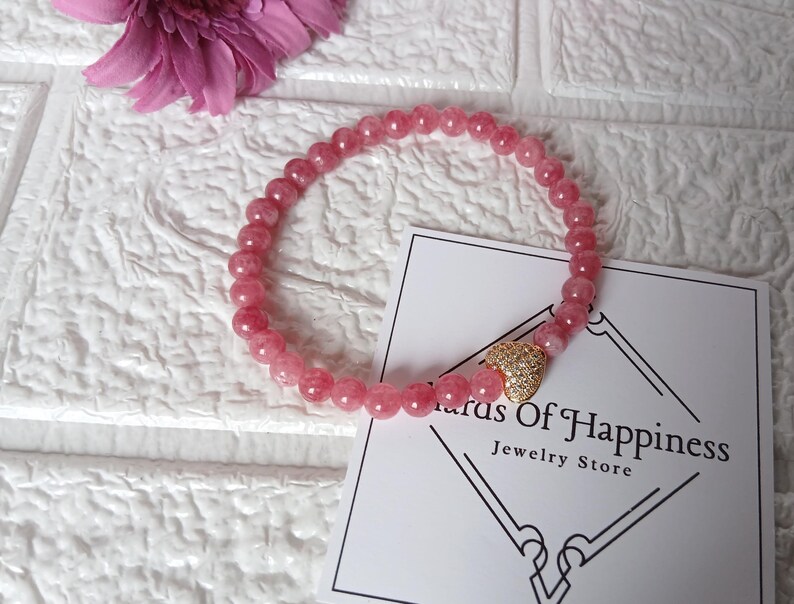 May include: A pink beaded bracelet with a gold heart charm. The bracelet is made of small, round, pink beads. The charm is gold and encrusted with small, clear stones. A card with the text "Status Of Happiness Jewelry Store" is also in the image.