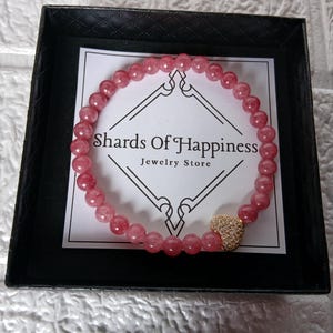 May include: A pink beaded bracelet with a gold heart charm, presented in a black box. The bracelet is made of small, round, pink beads. The heart charm is covered in small, sparkling stones. The box contains a card that reads "Shards Of Happiness Jewelry Store."