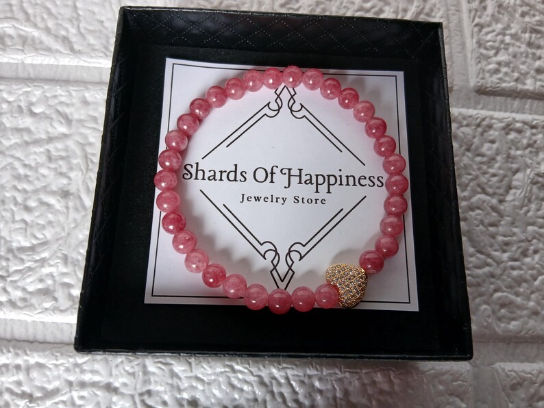 May include: A pink beaded bracelet with a gold heart charm, presented in a black box. The bracelet is made of small, round, pink beads. The heart charm is covered in small, sparkling stones. The box contains a card that reads "Shards Of Happiness Jewelry Store."