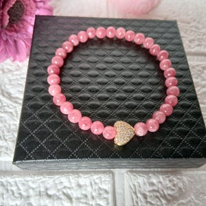 May include: A pink beaded bracelet with a gold heart-shaped charm, set with small, clear crystals. The bracelet is displayed on a black, textured box. A pink flower is visible in the background.