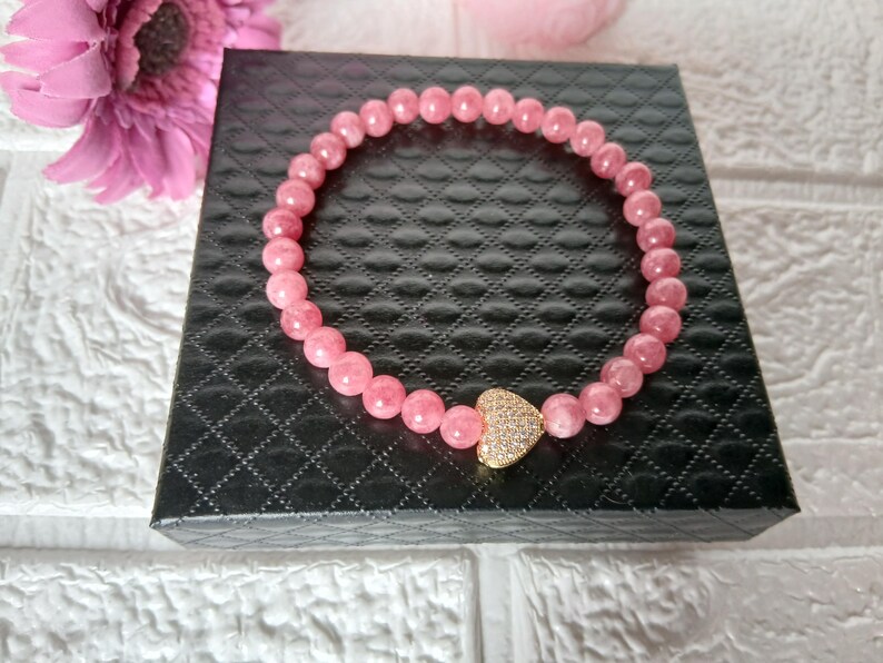 May include: A pink beaded bracelet with a gold heart-shaped charm, set with small, clear crystals. The bracelet is displayed on a black, textured box. A pink flower is visible in the background.