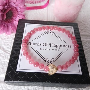 May include: A pink beaded bracelet with a gold heart charm rests on a black box with the text "Shards Of Happiness Jewelry Store". A pink flower and other pink objects are in the background. The bracelet is made of small, round, pink beads.