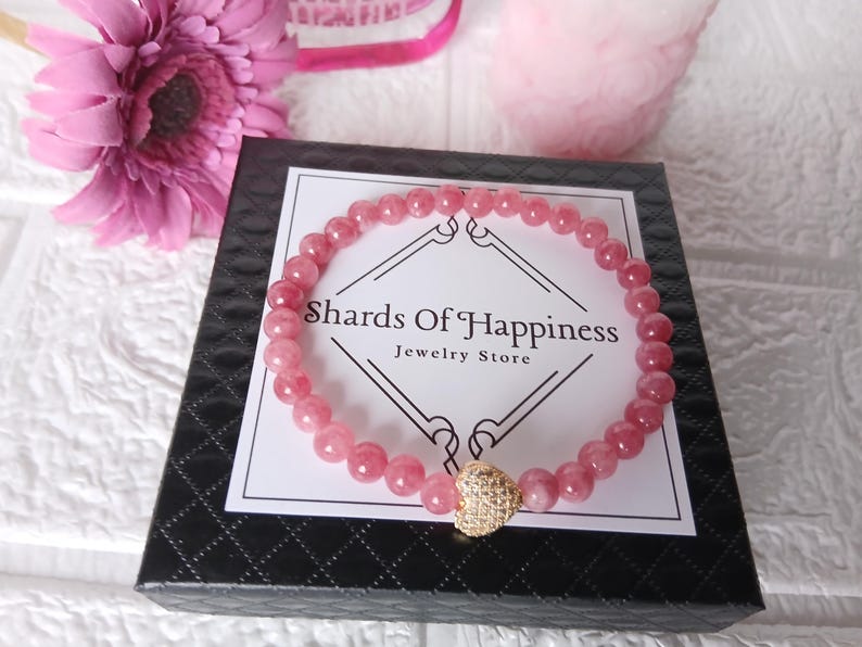 May include: A pink beaded bracelet with a gold heart charm rests on a black box with the text "Shards Of Happiness Jewelry Store". A pink flower and other pink objects are in the background. The bracelet is made of small, round, pink beads.