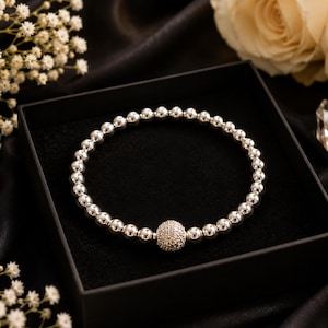 May include: A silver beaded bracelet featuring a single spherical charm encrusted with small clear stones. The bracelet is composed of polished metallic beads arranged in a circular pattern, centered around the textured, sparkling focal bead.