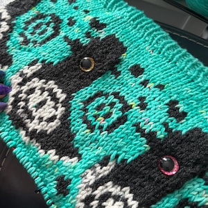 May include: Hand-knitted teal and black clutch with a geometric pattern. The clutch features a repeating design in black and white, with two decorative, gem-like embellishments. The knitted texture is visible, showcasing the craftsmanship of the handmade item.