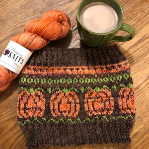 May include: A brown knitted hat with a pumpkin pattern. The hat has a band of orange, green, and brown knit around the bottom. The hat is sitting on a wooden surface with a cup of hot chocolate and a ball of orange yarn.