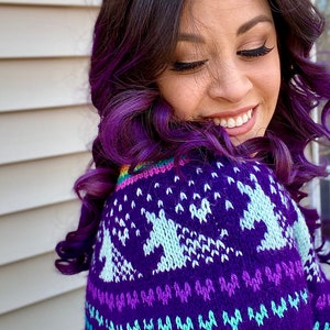 May include: A woman with purple hair is wearing a purple sweater with a white unicorn design. The sweater has a rainbow stripe across the shoulder.