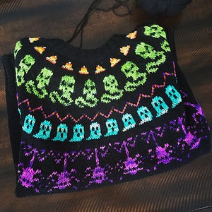 May include: Black knitted sweater with a colorful Halloween-themed design. The design features ghosts, skulls, and candy corn.