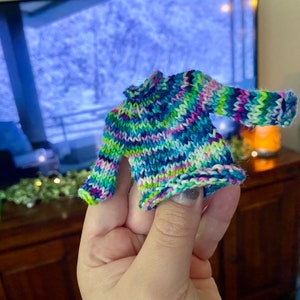 May include: A small, colorful knitted sweater with a striped pattern in shades of blue, green, pink, and white. The sweater has a turtleneck and long sleeves.