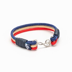 Op de afbeelding: Een gevlochten armband van donkerblauw, rood en beige touw met een zilveren vioolsleutelbedel.